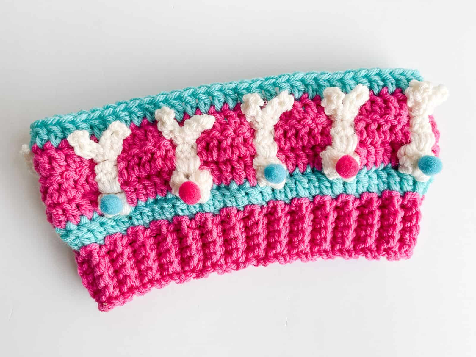 Free crochet bunny headband pattern in hot rose and turquoise with ivory raised bunny bobble stitches, shown as a flat lay with yarn and pom pom tails